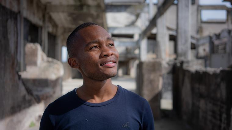 Beautiful News - Aphiwe Koti of Street Dogs Documentary standing thoughtfully with building ruins in background.