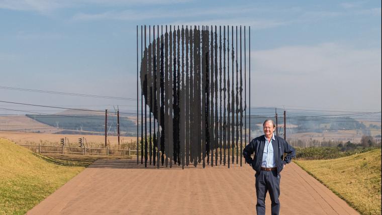 Man stands with his hands on his hips in front of a monument of Nelson Mandela