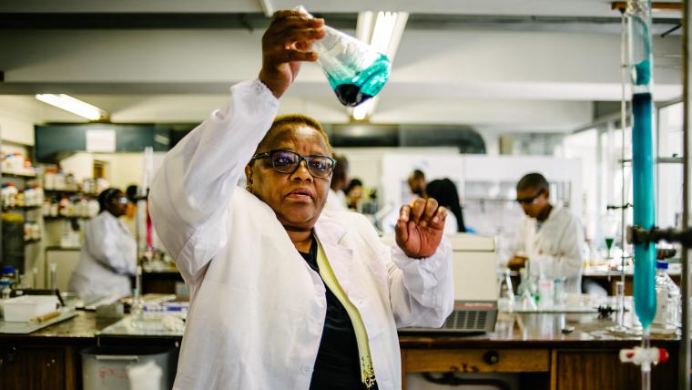 Black woman in a lab holds up a beaker containing liquid