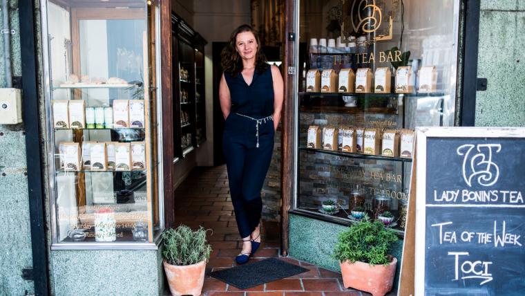 White woman standing by her tea shop door.