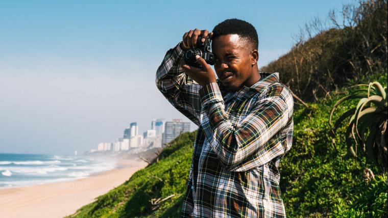 Young man stands on a beach holding a camera up to his eye